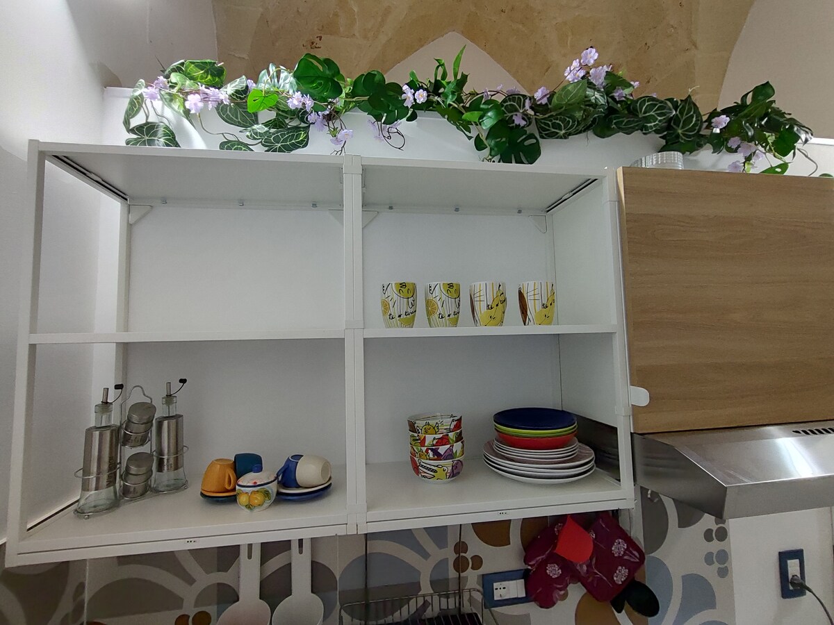 The kitchen is equipped with modern cabinetry featuring open shelving. Clear glass bottles and colorful dishware are arranged neatly, including cups and plates in various designs. A decorative vine accentuates the upper shelf, adding a touch of greenery to the space.