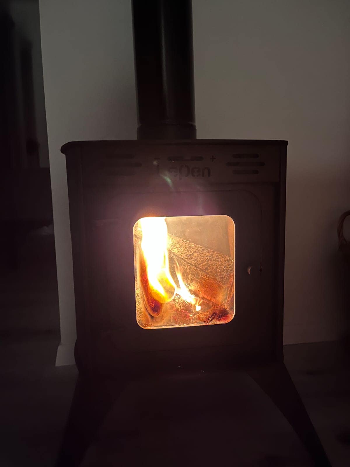 A wood stove is showcased, with a fire brightly burning inside, creating a warm glow. The stove's dark metal frame contrasts with the vibrant flames and logs visible through the glass front. The surrounding area is softly illuminated, enhancing the cozy ambiance.