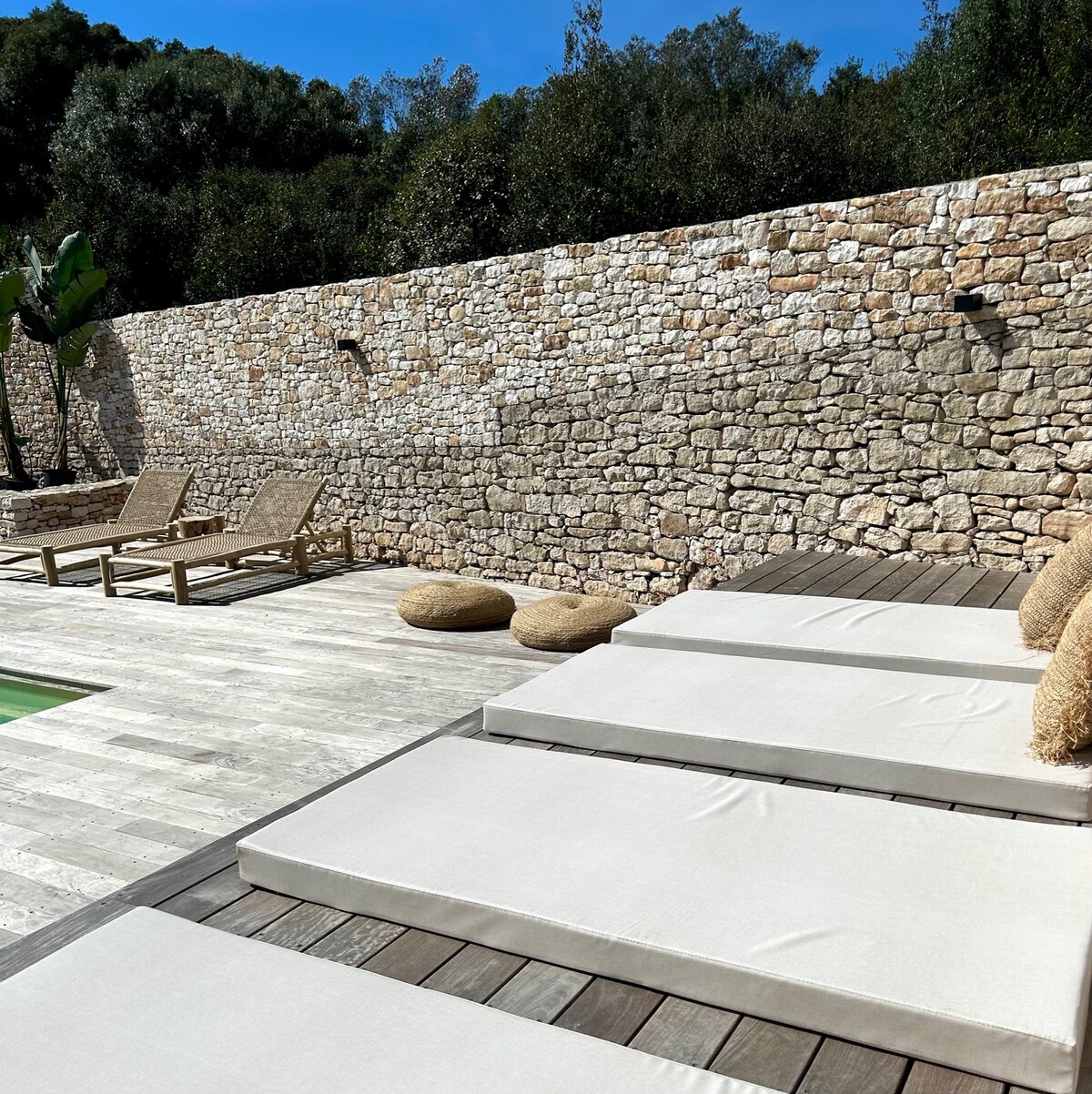 A serene outdoor space features a swimming pool surrounded by natural stone walls. Comfortable sun loungers are positioned on a wooden deck, alongside soft cushions. The tranquil atmosphere is enhanced by greenery and sunlight filtering through the trees.