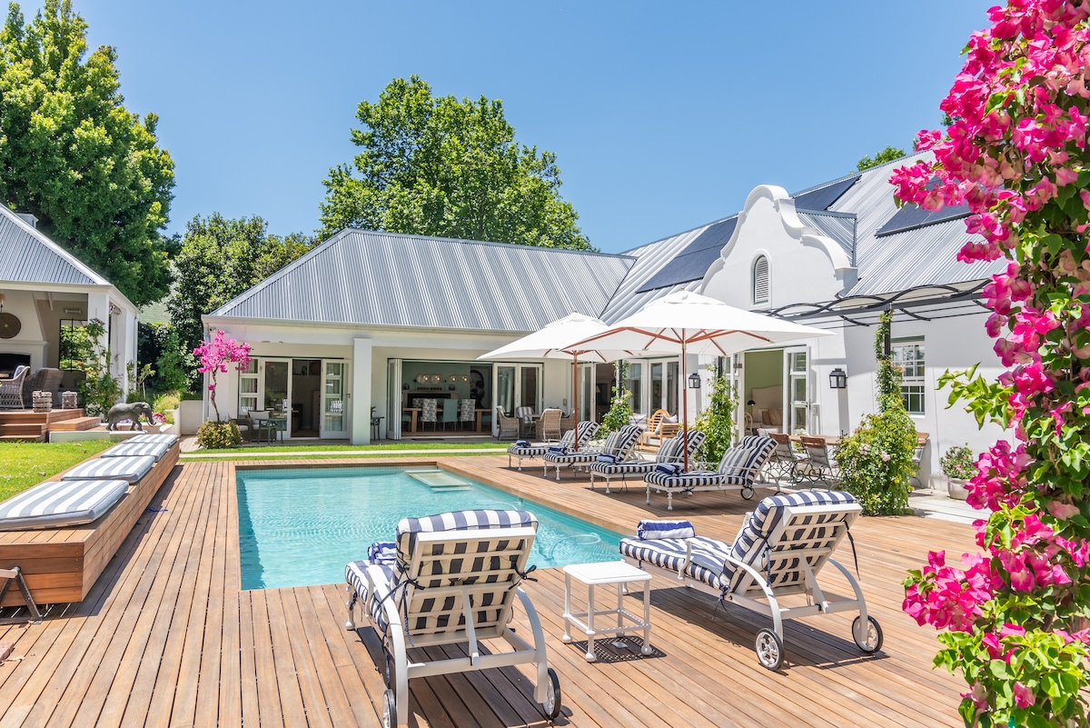 The outdoor area features a private heated swimming pool framed by a wooden deck. Comfortable sun loungers with striped cushions are positioned around the pool, and two umbrellas provide shade. Lush greenery and vibrant flowers create a serene atmosphere, enhancing the tranquil setting.