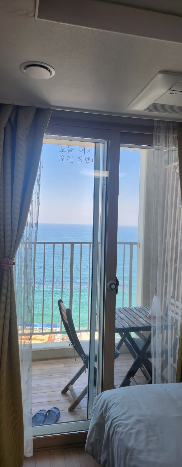 A sliding glass door offers a view of the ocean, with gentle waves visible in the distance. A small balcony features a table and two chairs, framed by sheer curtains that softly diffuse the natural light entering the room.