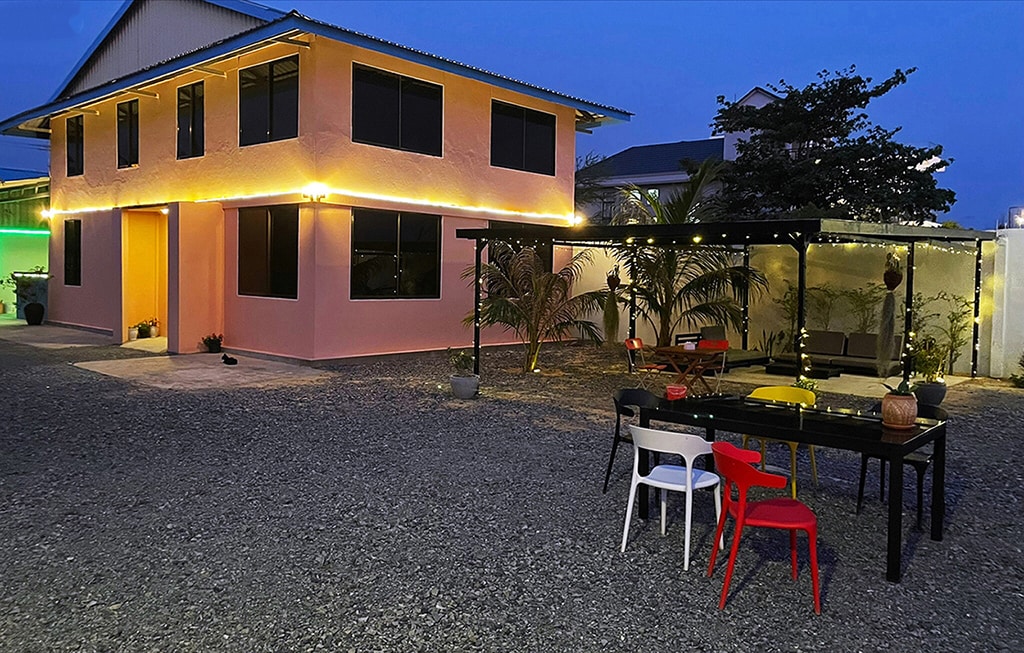 The exterior of the house is showcased at dusk, featuring a pink façade and large windows. String lights outline the structure, creating a welcoming glow. An outdoor dining area with multi-colored chairs and a dark table is surrounded by gravel and greenery, enhancing the inviting atmosphere.