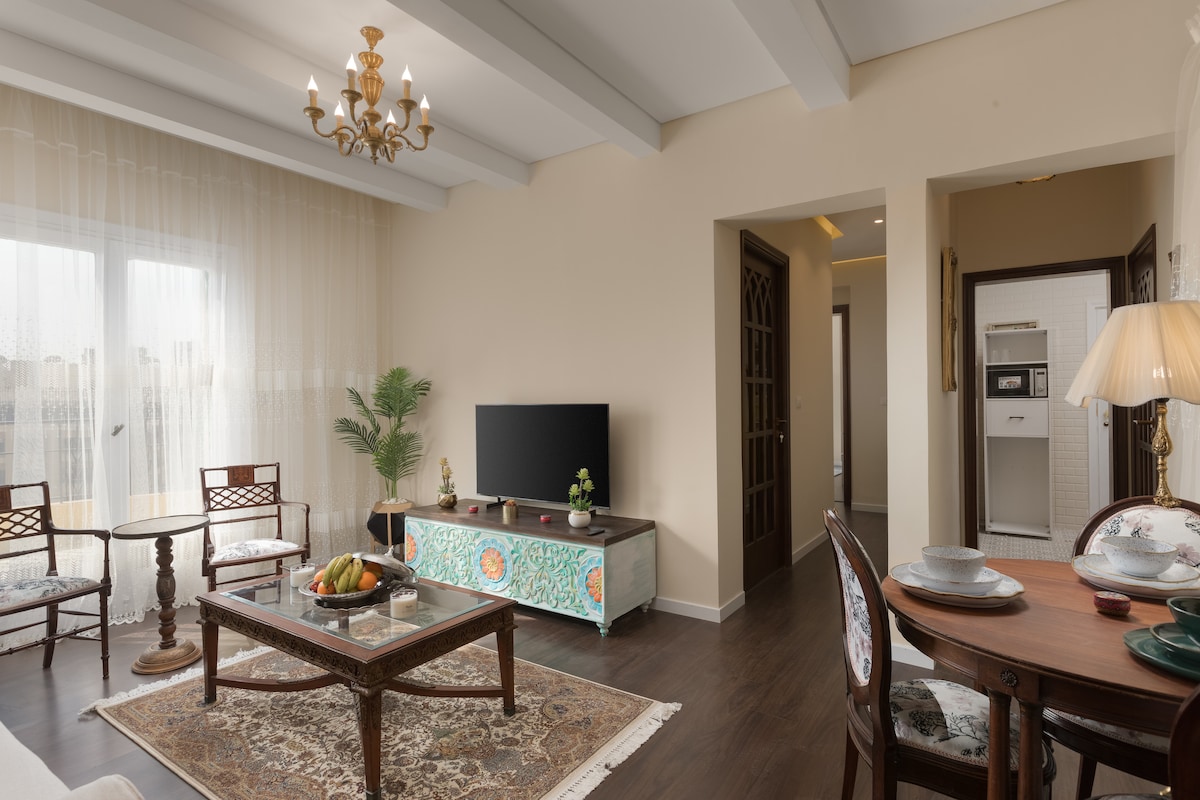 A spacious living area is complemented by a light, airy ambiance, featuring large windows with sheer curtains. A decorative chandelier hangs from the ceiling, while elegant furnishings include a wooden coffee table adorned with fruit, alongside a TV stand and dining area set for two.