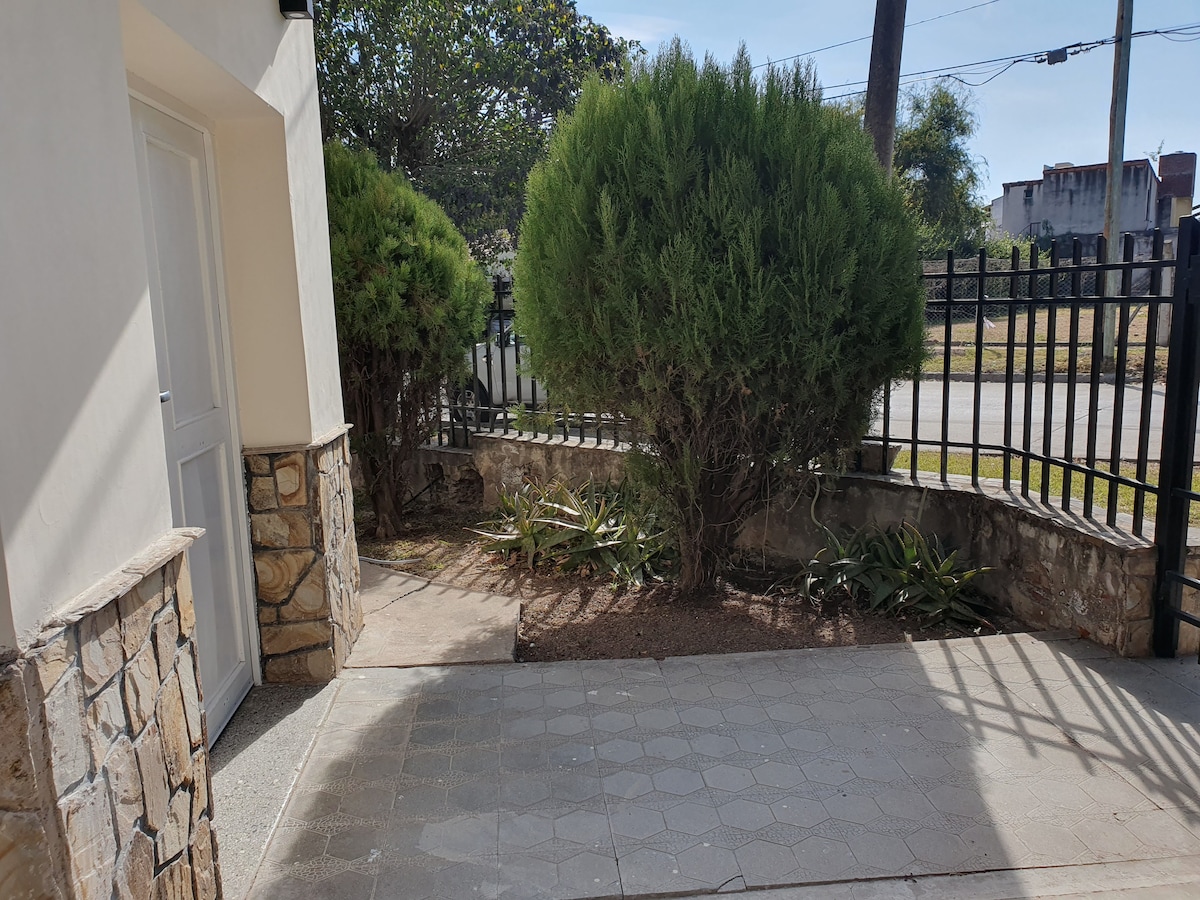 An exterior entrance is highlighted by well-maintained stone walls and a simple pathway. Lush greenery is present, including large shrubs and small plants, which provide a calming atmosphere. A black fence borders the space, offering a sense of privacy while allowing natural light to filter through.