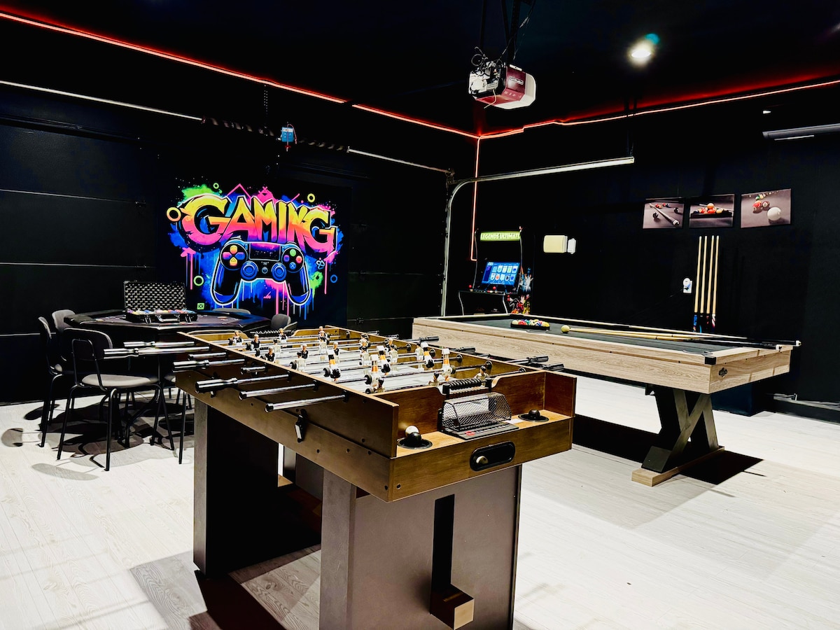 A game room showcases a variety of entertainment options, featuring a pool table on one side and a foosball table on the other. Bright neon 'Gaming' artwork adds a playful touch to the black walls, complemented by comfortable seating arranged for social interaction.