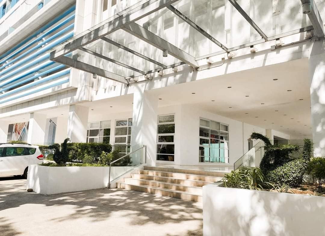 The entrance to the condominium features a modern design with large glass panels allowing natural light. A set of steps leads to the main entrance, framed by neatly arranged greenery and a parking area visible to the side.