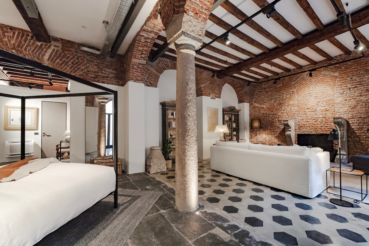 A spacious loft features exposed brick walls and rustic wooden beams. A large, four-poster bed is set in one area, while a comfortable seating arrangement is positioned nearby. The floor showcases unique hexagonal tiles, and a column stands prominently in the center of the room.