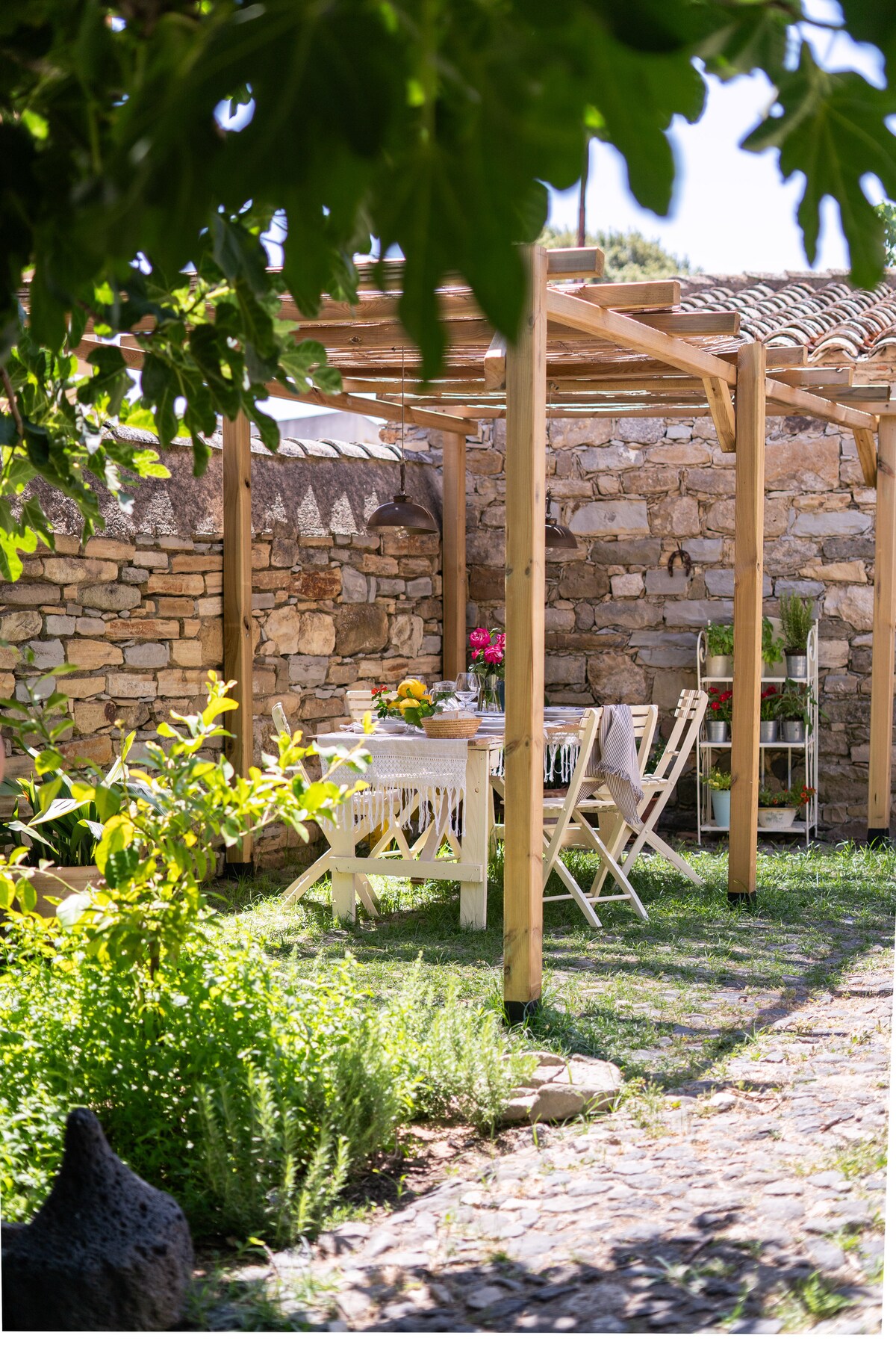 An outdoor dining area is set under a wooden pergola, surrounded by stone walls and lush greenery. A table is adorned with flowers and tableware, complemented by light-colored chairs. Sunlight filters through the leaves above, creating a peaceful and inviting atmosphere.