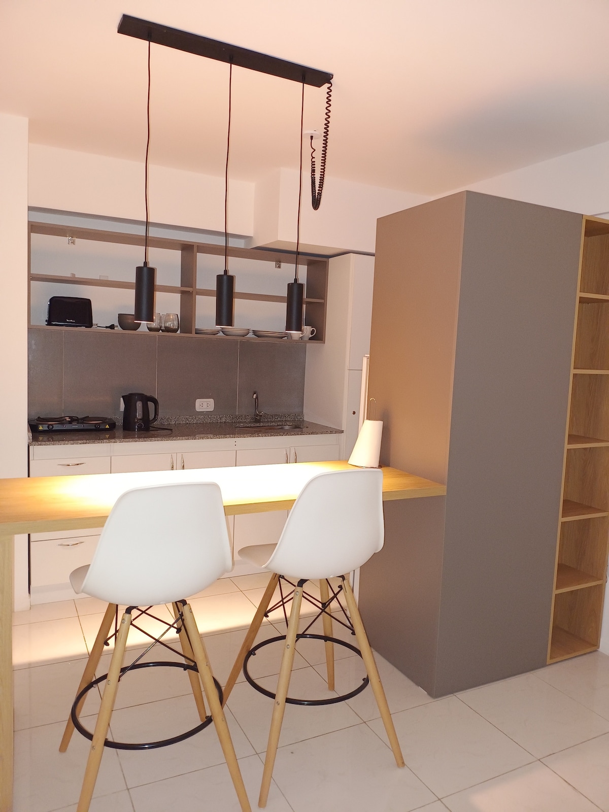 A modern kitchen area is highlighted by two white bar stools positioned at a wooden counter. Shelves are mounted above, displaying kitchenware. A sleek refrigerator stands nearby, with a neutral-toned cabinet providing additional storage. Warm lighting enhances the inviting atmosphere of the space.