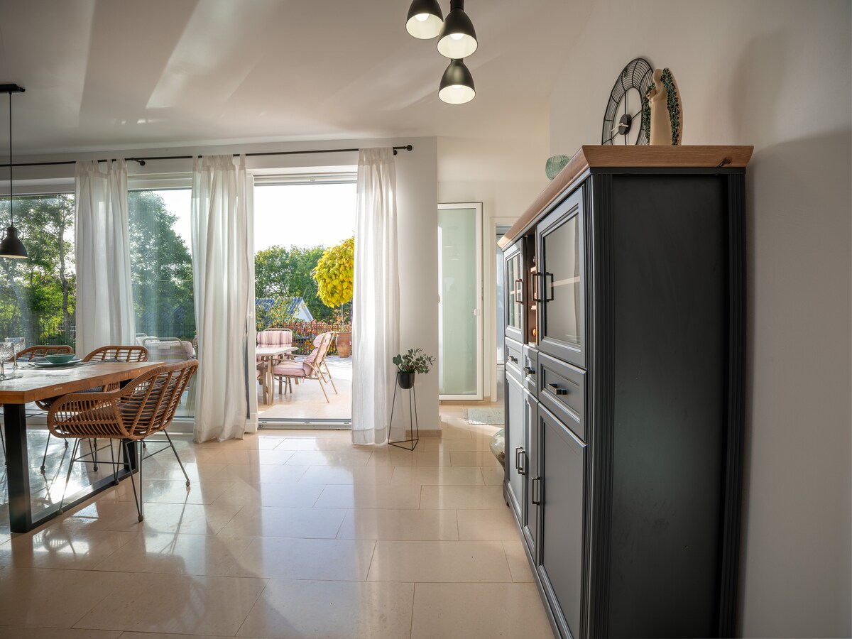 A spacious dining area is highlighted by a round table surrounded by woven chairs. Large glass doors allow natural light to fill the space, leading to an outdoor seating area. A stylish cabinet adds a touch of charm and functionality to the room.