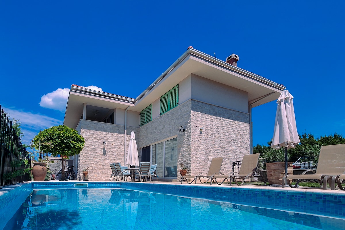 A modern villa is presented, featuring a private pool with crystal clear water. Sun loungers are arranged around the pool area, and shaded umbrellas provide relief from the sun. The building showcases a light-colored facade and large windows, reflecting the serene outdoor surroundings.