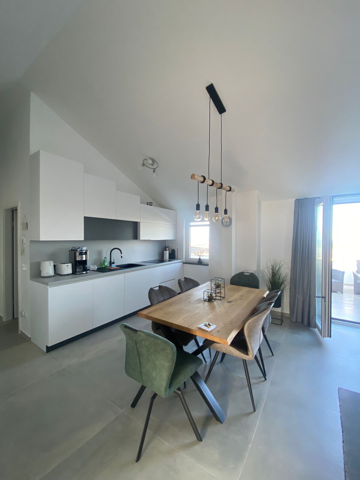 A modern kitchen area features sleek cabinetry and stainless steel appliances, including a coffee maker and sink. A wooden dining table with six chairs is positioned next to a large window, allowing natural light to fill the space. A contemporary light fixture hangs above the table.