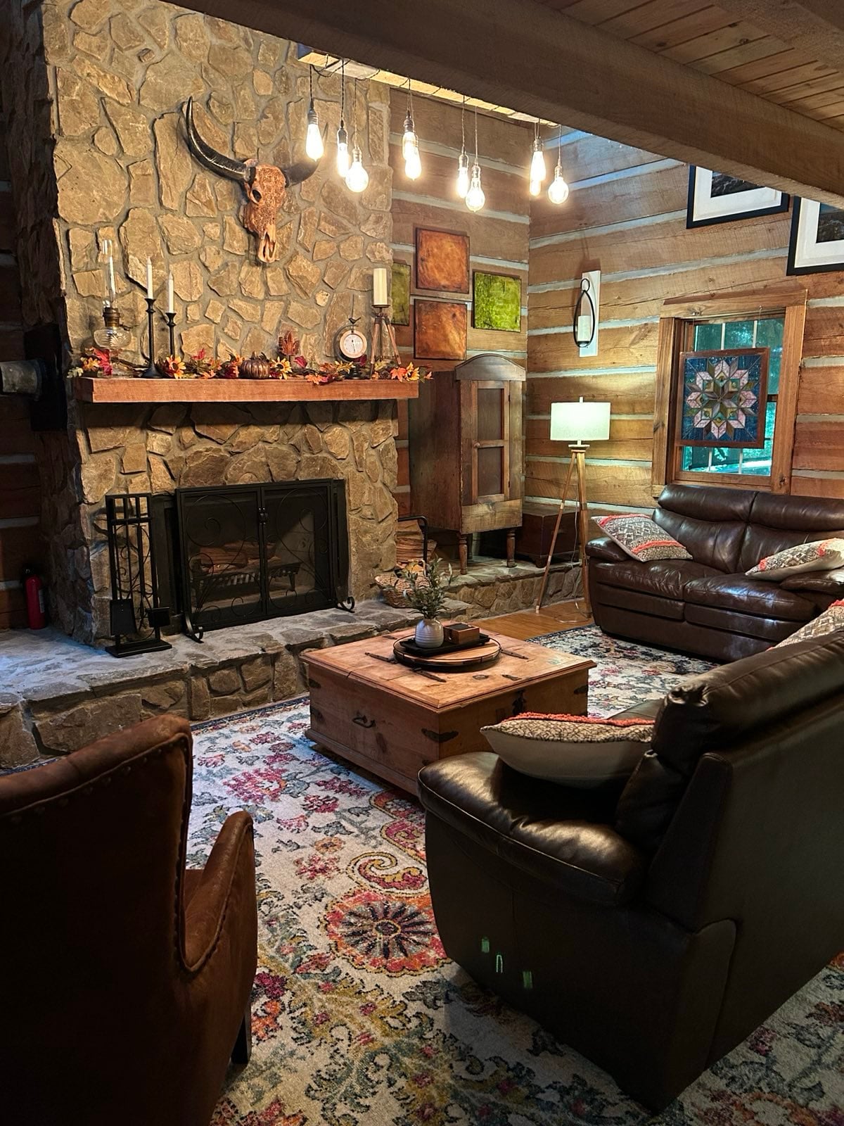 A rustic living area features a stone fireplace with wood accents, adorned with decorative objects and artwork. Comfortable leather seating surrounds a central coffee table atop a colorful area rug. Warm lighting creates a cozy ambiance in the wooden interior.