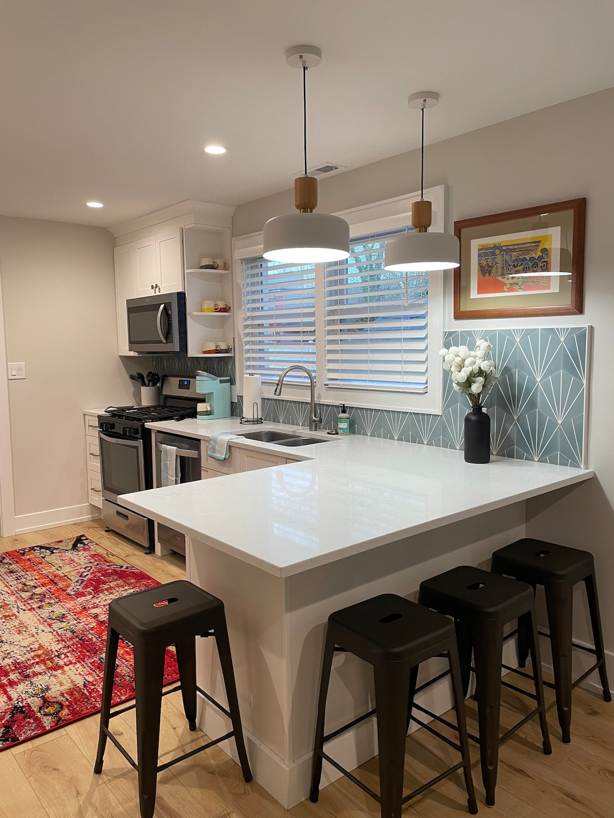 A modern kitchen featuring white cabinetry and a light countertop. A black microwave and a stainless steel stove are visible. Four metal bar stools are positioned at the counter. Natural light filters through the blinds, illuminating a decorative vase with flowers on the counter.