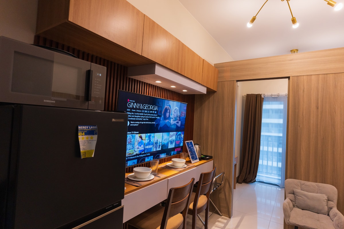 A modern kitchen area features wood-paneled cabinetry and sleek appliances. A large television is mounted on the wall, displaying a streaming service. The dining table is set for two, accompanied by stylish chairs. A light-filled balcony door partially opens to invite natural light.