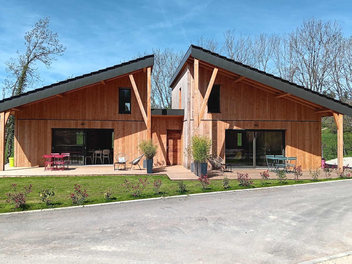 The exterior of the eco-friendly duplex home showcases wooden façades and a modern design. Large glass windows reflect natural light, while the landscaped garden features greenery and flower beds. A pathway leads to the entrance, creating an inviting approach to the accommodation.
