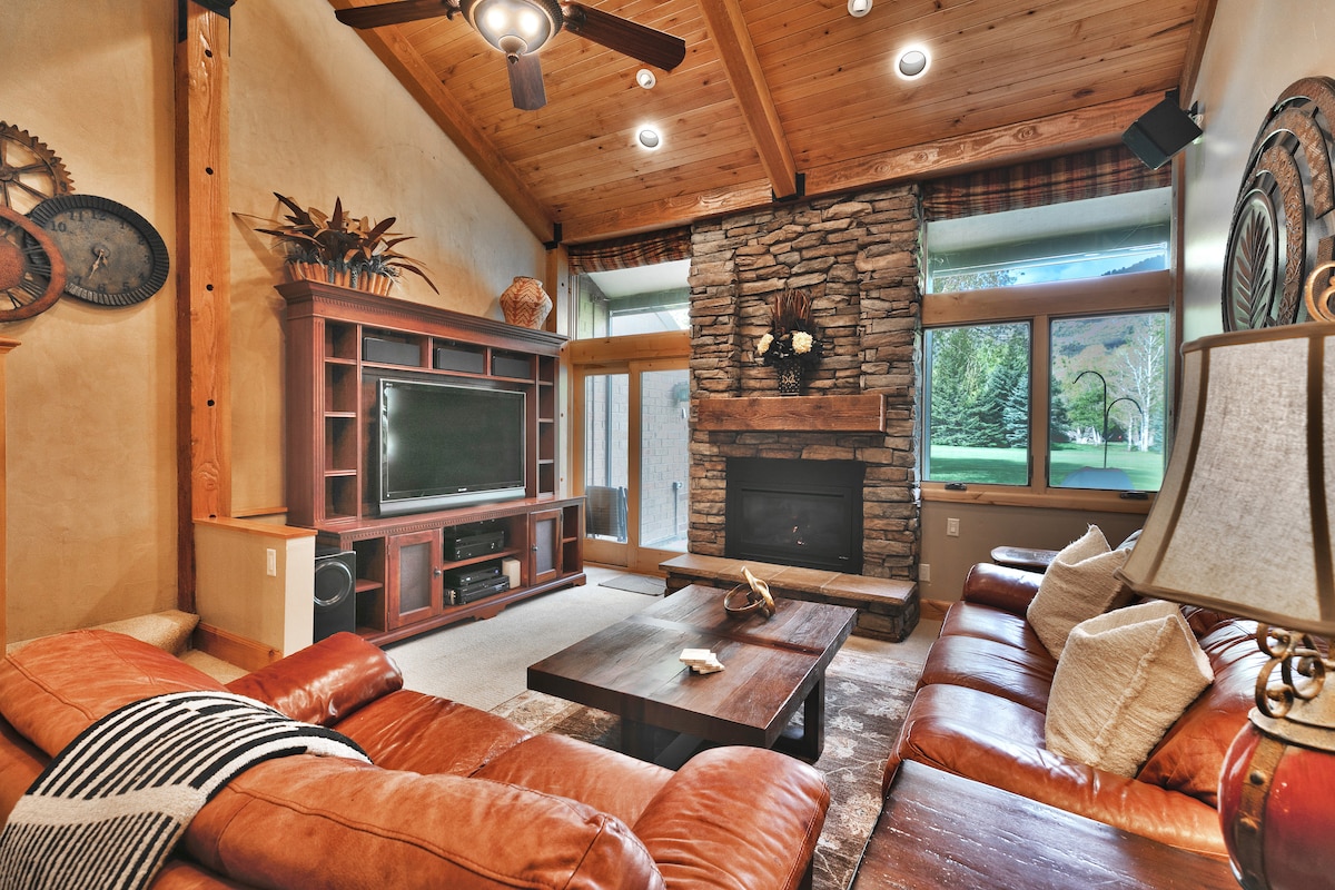 A spacious living area features a stone fireplace and large windows offering views of the outdoor landscape. Comfortable leather sofas surround a wooden coffee table, while a flat-screen TV is mounted above an entertainment unit. Natural wood beams enhance the cozy ambiance.