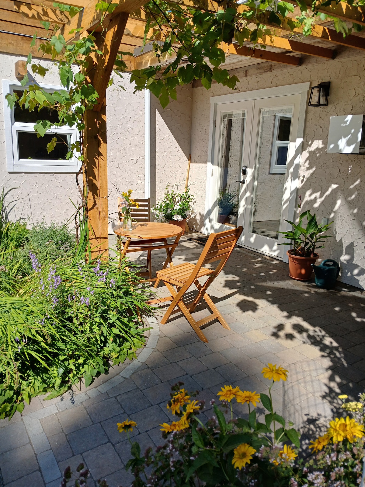 A charming patio area is highlighted by a wooden table and two folding chairs, surrounded by vibrant greenery and flowers. Natural light filters through a pergola overhead, creating a serene outdoor space ideal for relaxation.