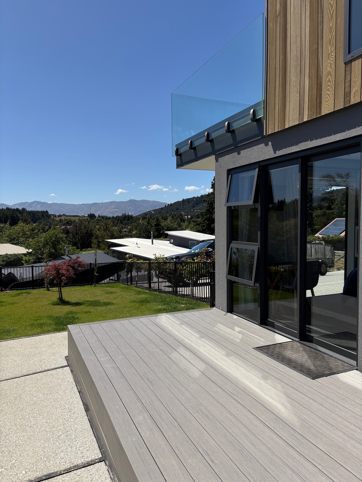 The exterior view showcases a deck area with wooden planks leading to expansive glass doors. A landscape of green hills and mountains is visible in the background, accented by clear blue skies and sunlight reflecting off the surfaces.