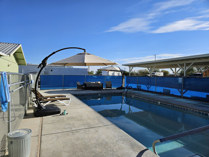 A clear swimming pool is surrounded by a concrete deck, featuring a shaded lounging area with a large umbrella and outdoor seating. The space is enclosed with blue fencing, providing a private and serene atmosphere under a sunny sky.
