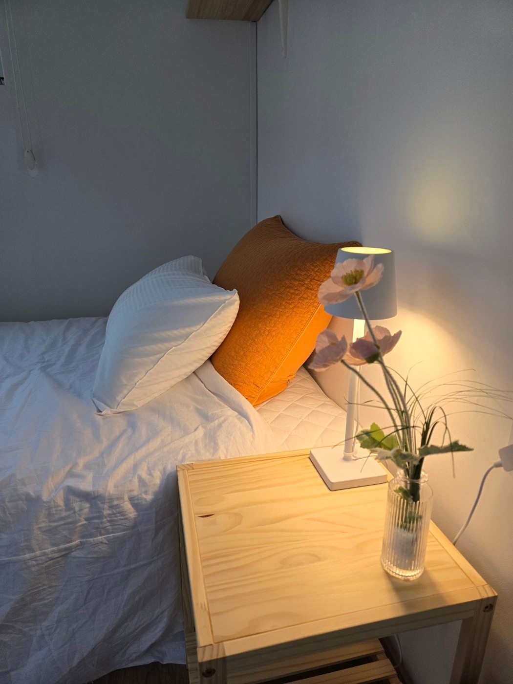 A bright sleeping area is presented, featuring a spacious bed adorned with crisp white linens and a textured orange pillow. A simple bedside table holds a slender vase with delicate flowers, complemented by a soft lamp that provides warm light.