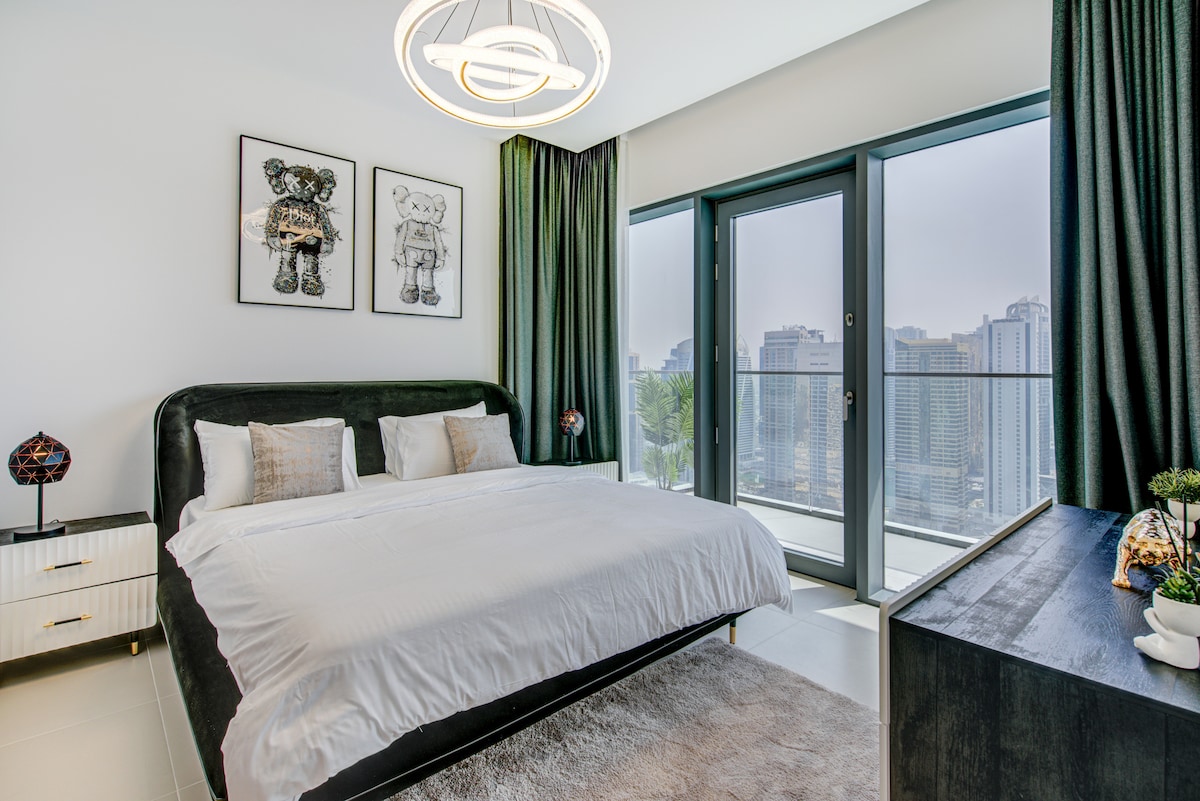 A spacious bedroom is presented with a large bed featuring a dark upholstered headboard. Accented by two bedside lamps, it is illuminated by natural light coming through sliding glass doors leading to a balcony, which offers views of the city skyline.