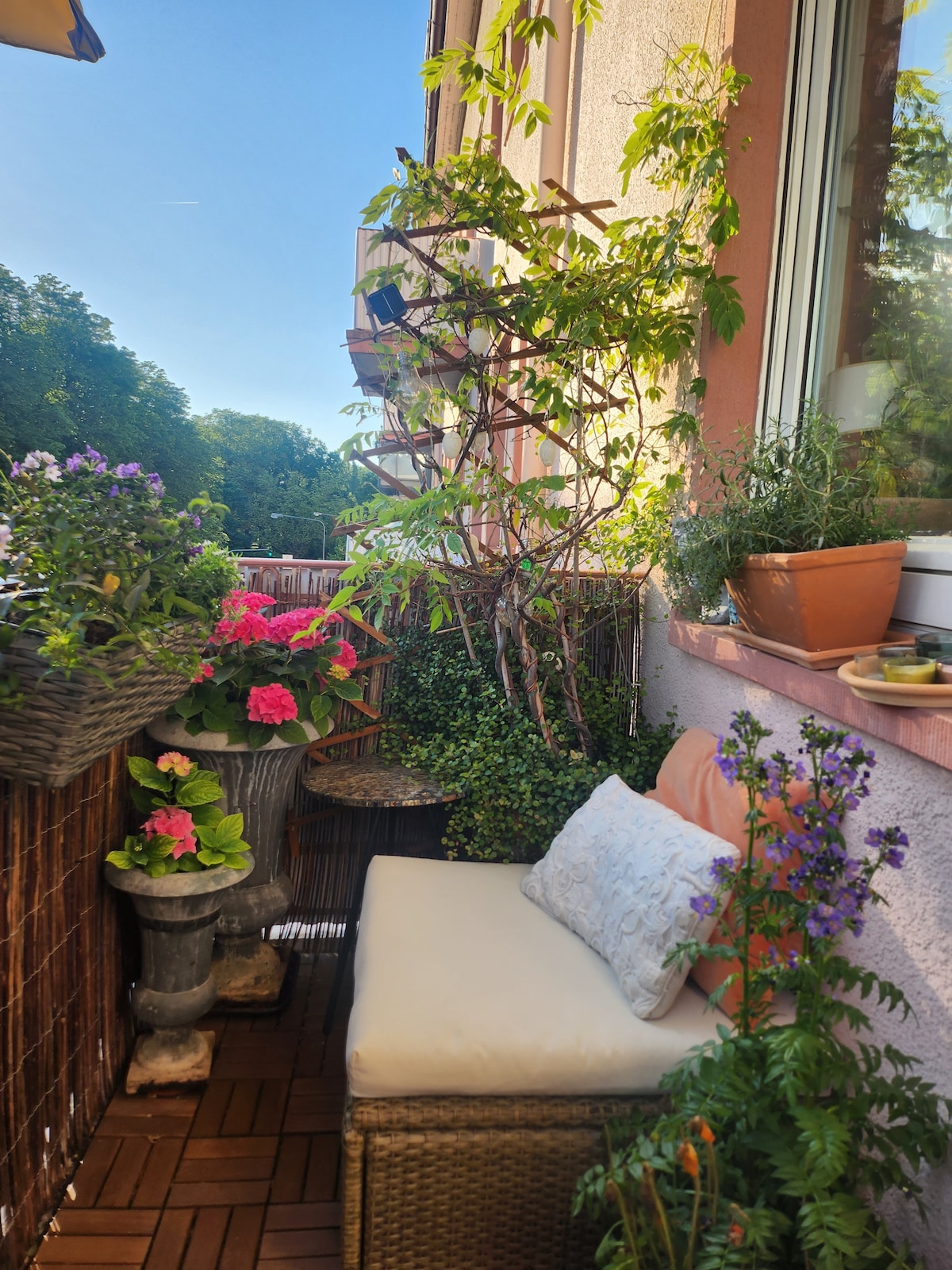 A small balcony features a cozy seating area with a cushion-covered chair. Potted plants and flowers in vibrant colors create a lush environment, while trellises support climbing greenery. Natural light bathes the space, enhancing the relaxing atmosphere for morning coffee or evening relaxation.