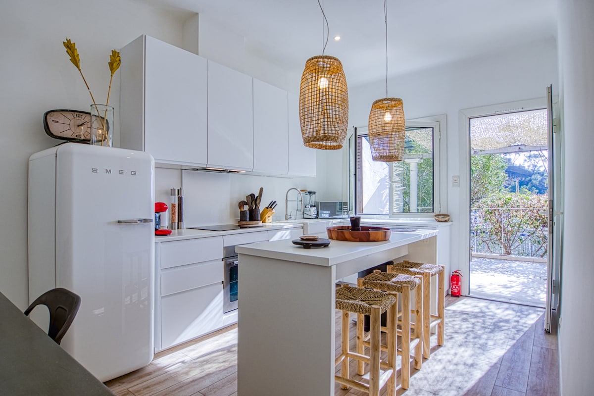 A modern kitchen features sleek white cabinetry and a large central island with high stools. Natural light filters through open doors leading to the outside. Rattan pendant lights hang above, and a vintage-style refrigerator adds character to the space.