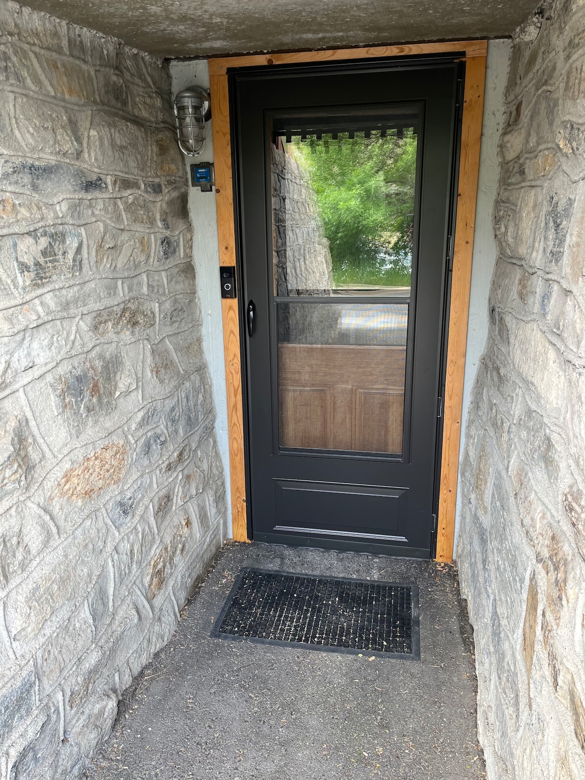 A well-constructed entrance features a black screen door framed by natural stone walls. A small mat is positioned in front of the door, and natural greenery is visible beyond the entrance, suggesting an inviting approach to the rental.