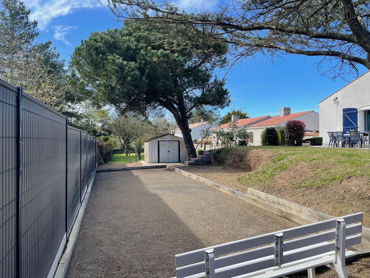 A private petanque court is set within a spacious outdoor area, bordered by lush greenery and trees. A small shed is visible in the background, alongside an upward slope leading to a well-maintained lawn. Clear blue skies enhance the openness of the space.