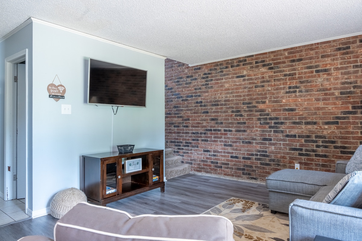 A living area showcases a combination of light-colored walls and exposed brick accents. A television is mounted on the wall opposite a gray sectional sofa. A decorative area rug can be seen on the floor, adding texture to the inviting space.