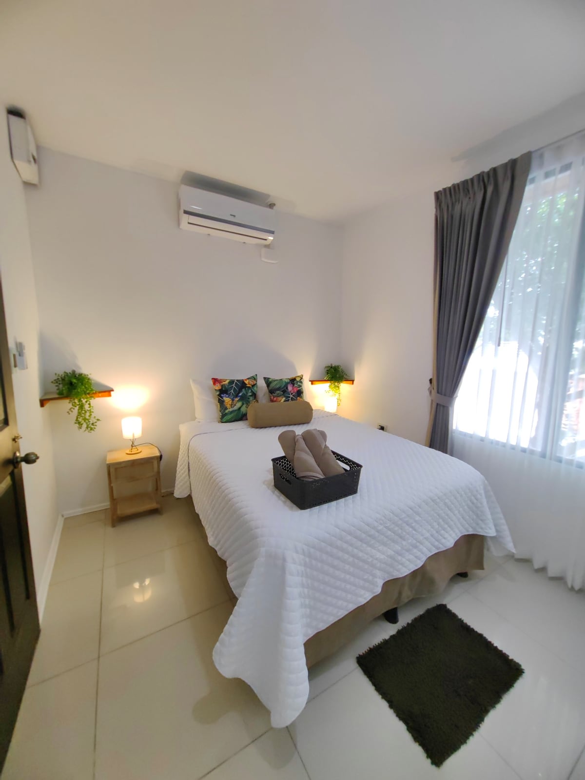 A cozy bedroom features a spacious bed with a light quilt, two bedside lamps, and decorative plants on each side. Natural light is allowed through sheer curtains, creating a bright and airy ambiance. An air conditioning unit is mounted above the bed for comfort.