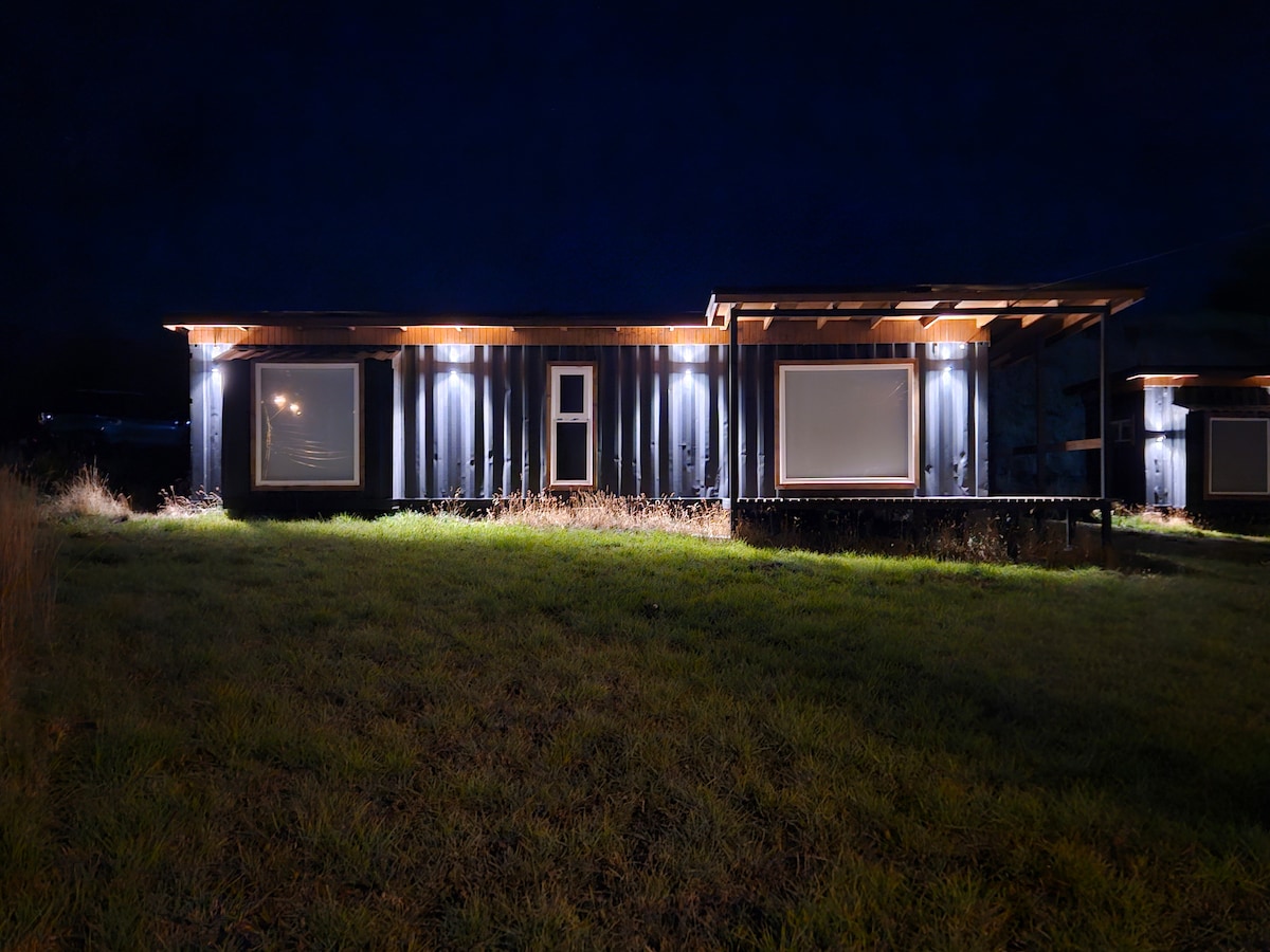 The exterior of the tiny house is illuminated at night, revealing a modern design with sleek lines. Outdoor lighting highlights the container structure's features, including large windows and a covered porch. The surrounding grassy area enhances the sense of seclusion and tranquility.