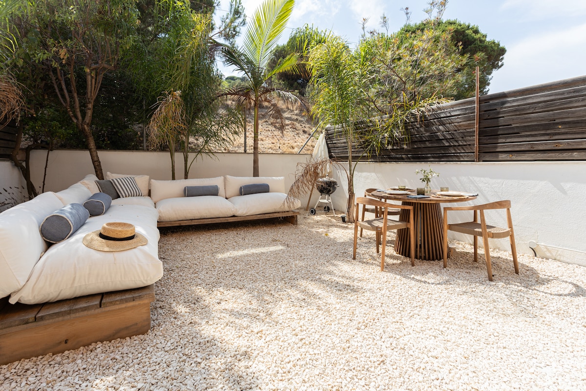 An outdoor terrace is presented with a cozy seating area featuring a spacious white sectional sofa with decorative pillows. A wooden dining table accompanied by four chairs sits nearby, surrounded by gravel and natural greenery, creating a relaxed atmosphere for gatherings.