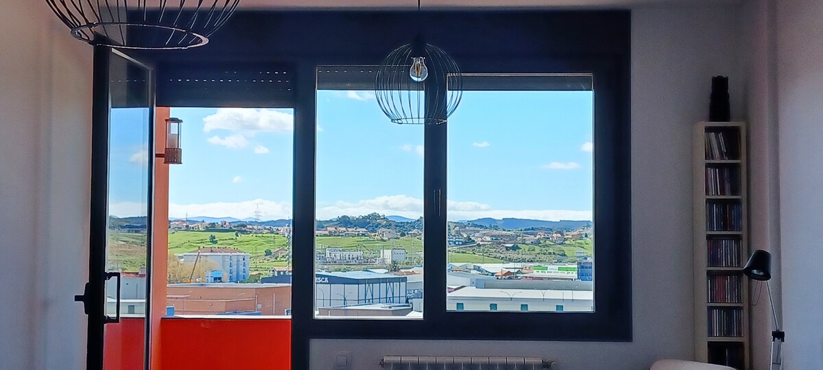 Large windows provide a view of rolling hills and distant mountains. The landscape features varying shades of green and blue under a clear sky, with scattered clouds. A modern light fixture adds character near the window, creating a bright and airy feel in the space.