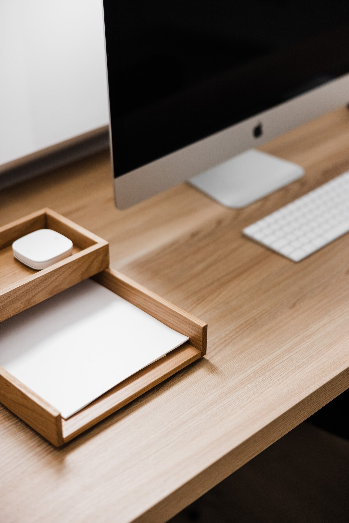 A minimalist workspace is displayed, featuring a sleek computer monitor positioned on a smooth wooden desk. A wooden organizer holds a notepad and a wireless device, while a compact keyboard adds to the clean design.