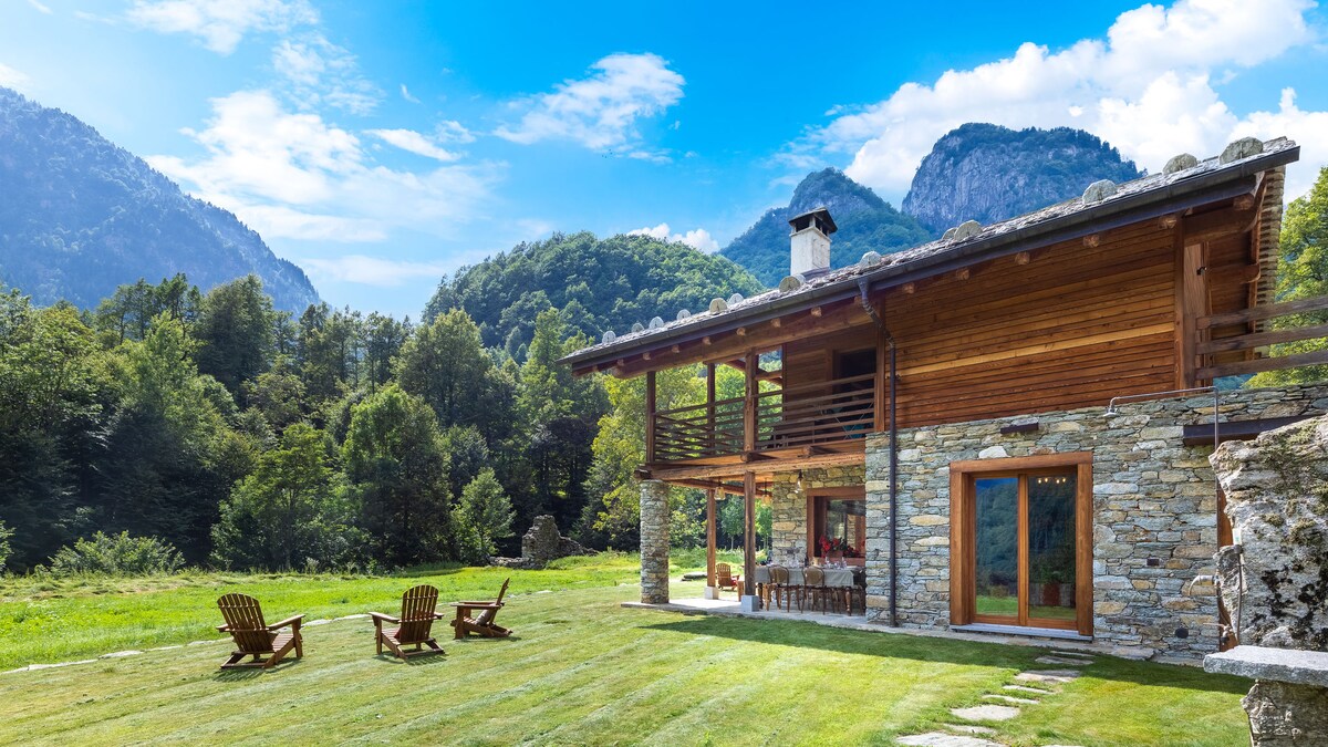 A charming chalet structure showcases a combination of wooden and stone elements. The surrounding garden features a well-maintained lawn with several outdoor seating chairs, framed by trees and mountains in the background, providing a serene natural setting.