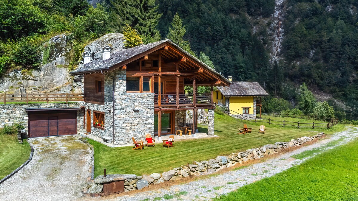 The exterior of the chalet is showcased, featuring a rustic stone facade and wooden balcony. A well-maintained lawn surrounds the property, with wooden deck chairs placed on the grass. The entrance is accessed via a gravel path, leading to a dual-door garage on the left.