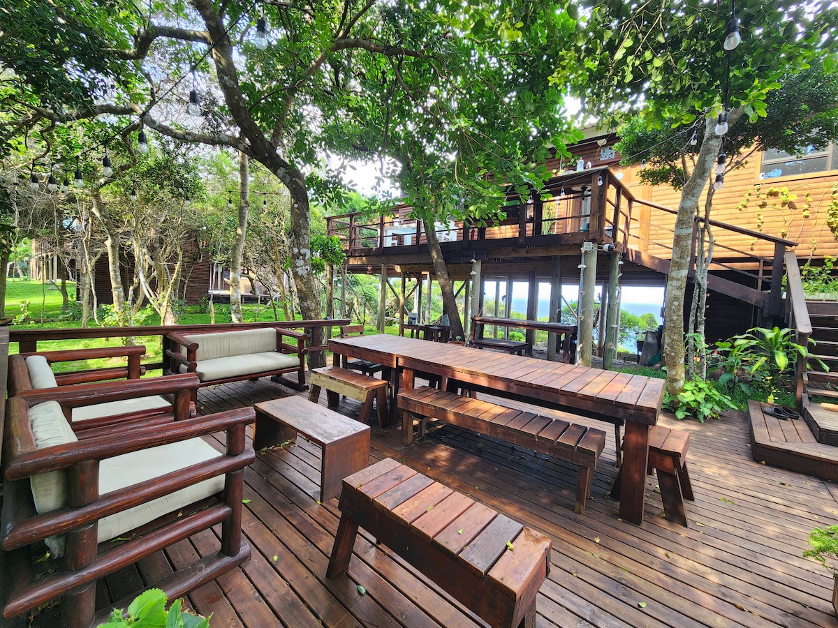 An outdoor gathering space is presented, featuring wooden furniture arranged among lush greenery. Several benches and tables are situated on a wooden deck, providing a natural atmosphere. A raised wooden structure is visible in the background, blending harmoniously with the surrounding trees.