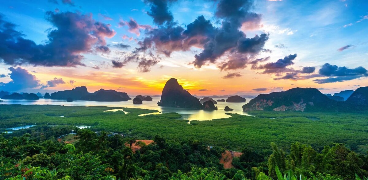 A panoramic view of lush green islands and tranquil waters is presented at sunset, with soft hues of orange, pink, and blue blending in the sky. The horizon showcases a variety of mountainous silhouettes, reflecting the serene beauty of the surrounding nature.