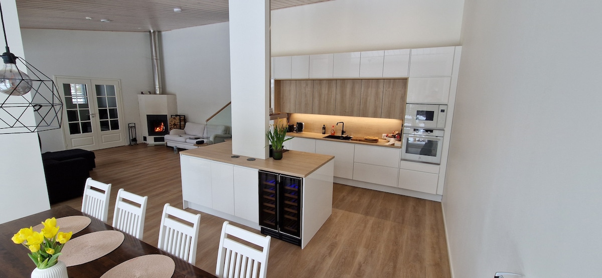 A modern kitchen is showcased with sleek cabinetry and a central island, equipped with a wine cooler. A dining table with white chairs is visible, adorned with a vase of yellow flowers. In the background, a cozy living area features a fireplace.