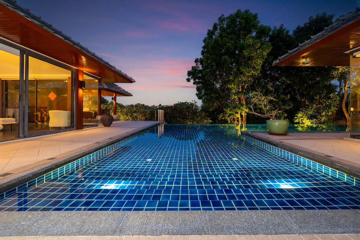 An infinity pool is set against a backdrop of lush greenery, with vibrant blue tiles reflecting the evening sky. Soft lighting is embedded in the pool, enhancing the tranquil ambiance, while modern villa structures frame the scene, showcasing an open and inviting layout.