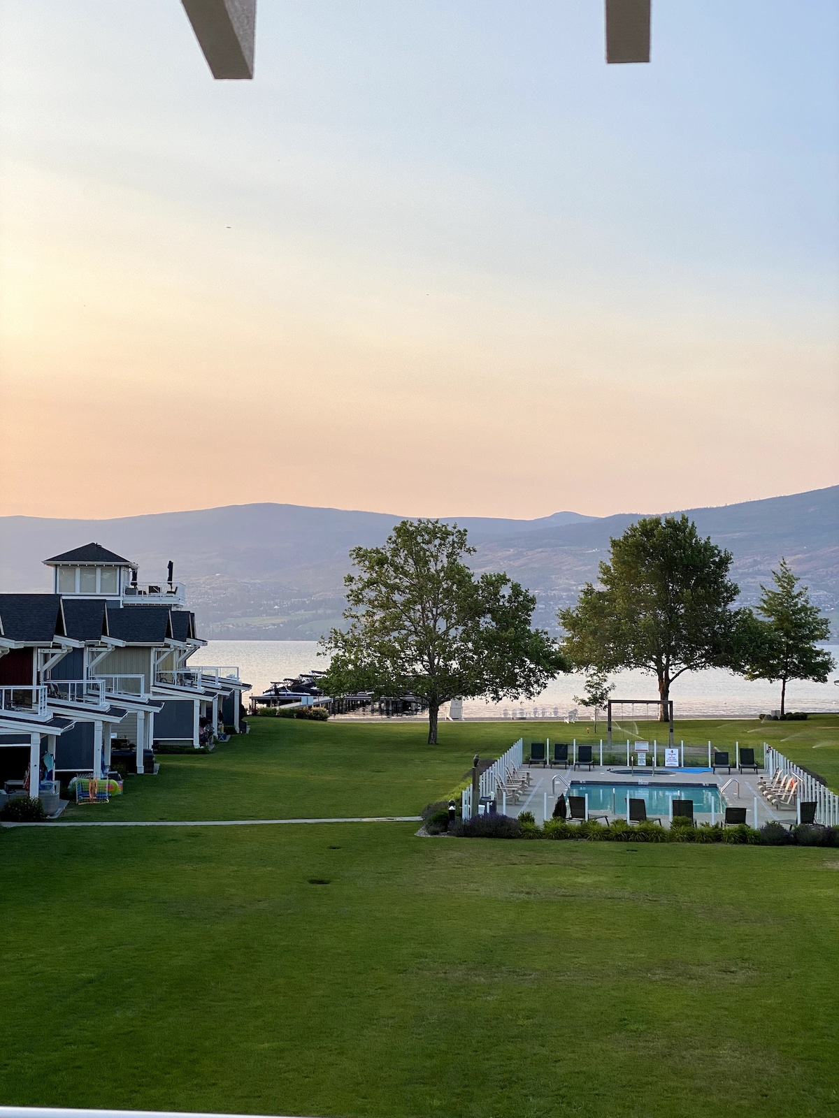 A serene view captures the spacious grassy area adjacent to the cottages, with a heated pool and hot tub in the foreground. The calm waters of Okanagan Lake and distant mountains create a gentle backdrop, highlighting the tranquility of this lakeside getaway.