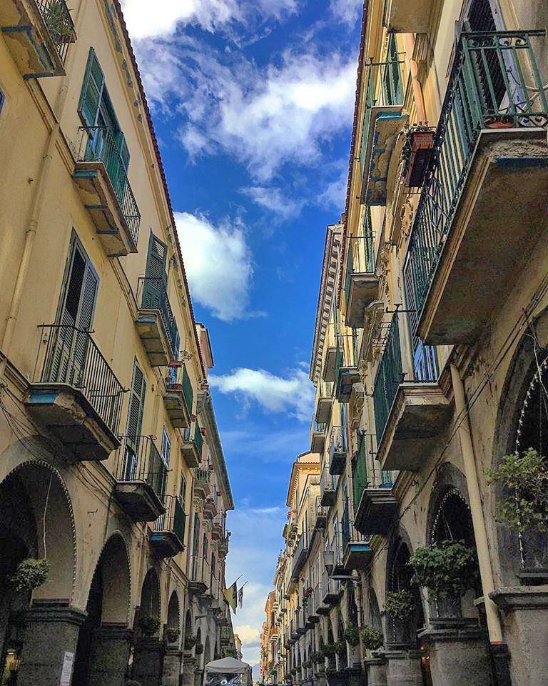 A view of a charming historic street is captured, featuring tall buildings adorned with balconies. The sky is highlighted by scattered clouds, creating a bright contrast against the architecture. Arched columns line the walkway, inviting exploration of the vibrant atmosphere.