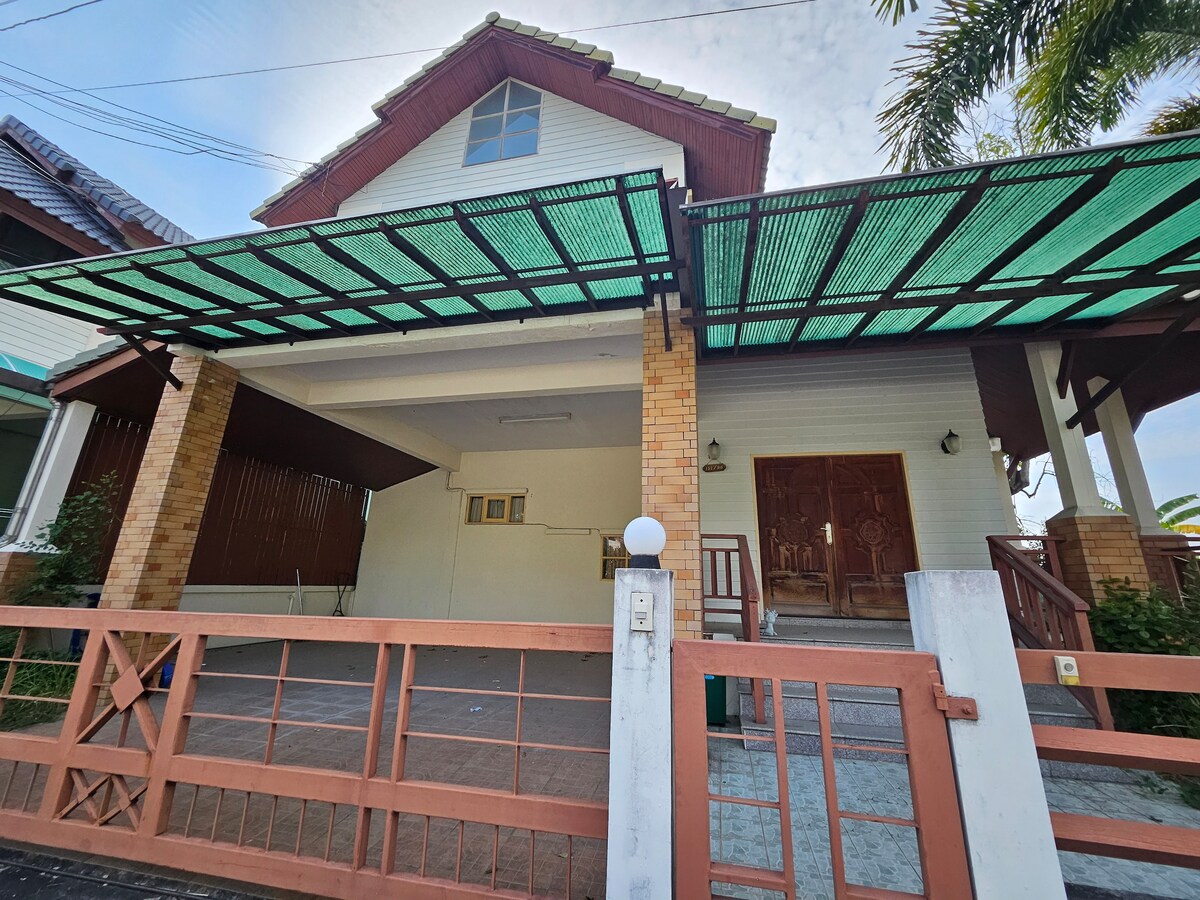 The exterior of a two-story house features a covered entrance, providing shade over the front space. Brick accents complement the wooden door, while surrounding greenery enhances the home’s natural setting. A paved area leads to the entrance, creating a welcoming approach.