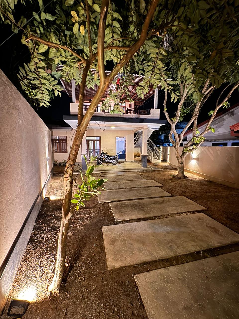 A well-lit pathway leads to the entrance of the villa, framed by trees and bordered by landscaped gravel. The structure features a welcoming facade, with outdoor stairs leading to a balcony above, while a parked motorcycle adds a touch of convenience.