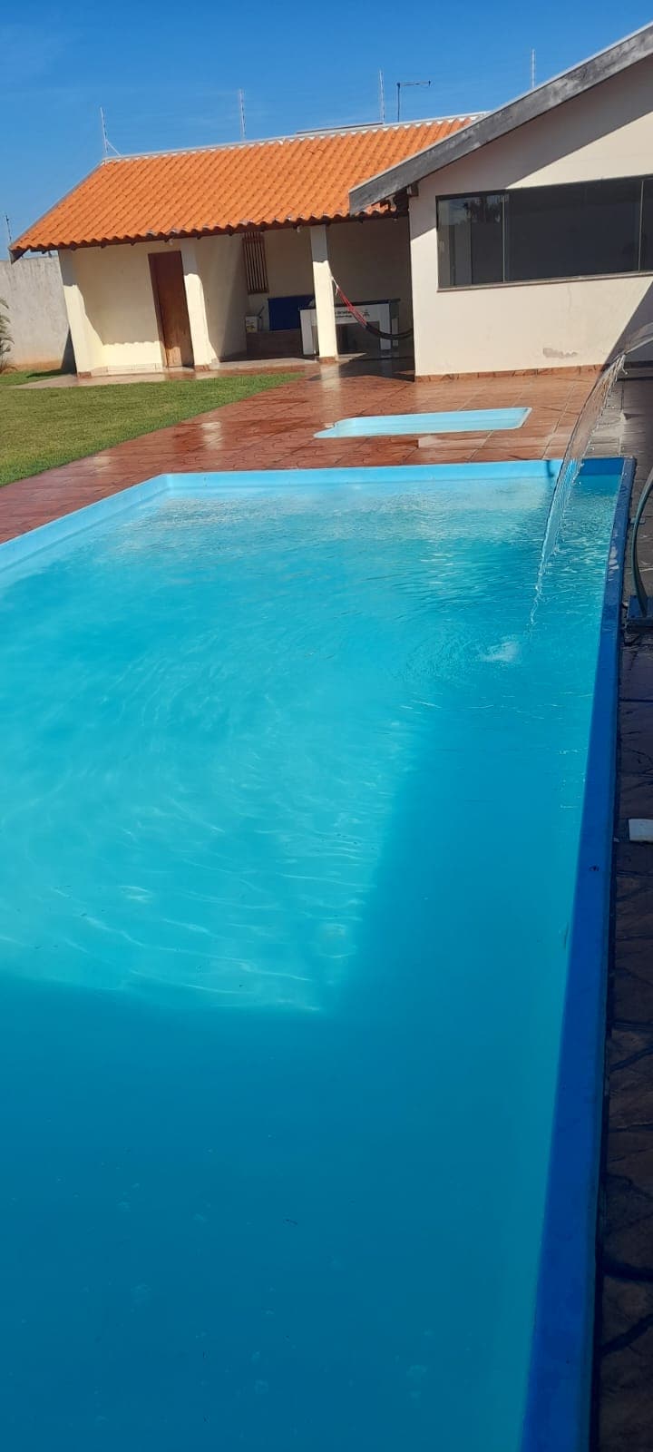 A clear swimming pool is visible, with water reflecting sunlight. Surrounding areas feature a tiled deck and green grass, creating an open and inviting outdoor space. A gentle water feature is seen along the pool's edge, adding to the serene environment.