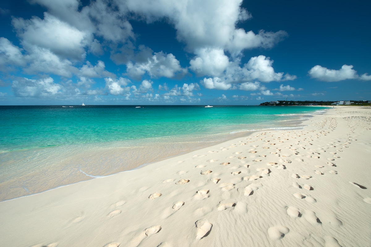 A tranquil beach scene features soft, white sand leading to clear, turquoise waters. Gentle waves lap at the shore, and a few boats can be seen in the distance. The sky is adorned with scattered clouds, creating a serene coastal atmosphere.