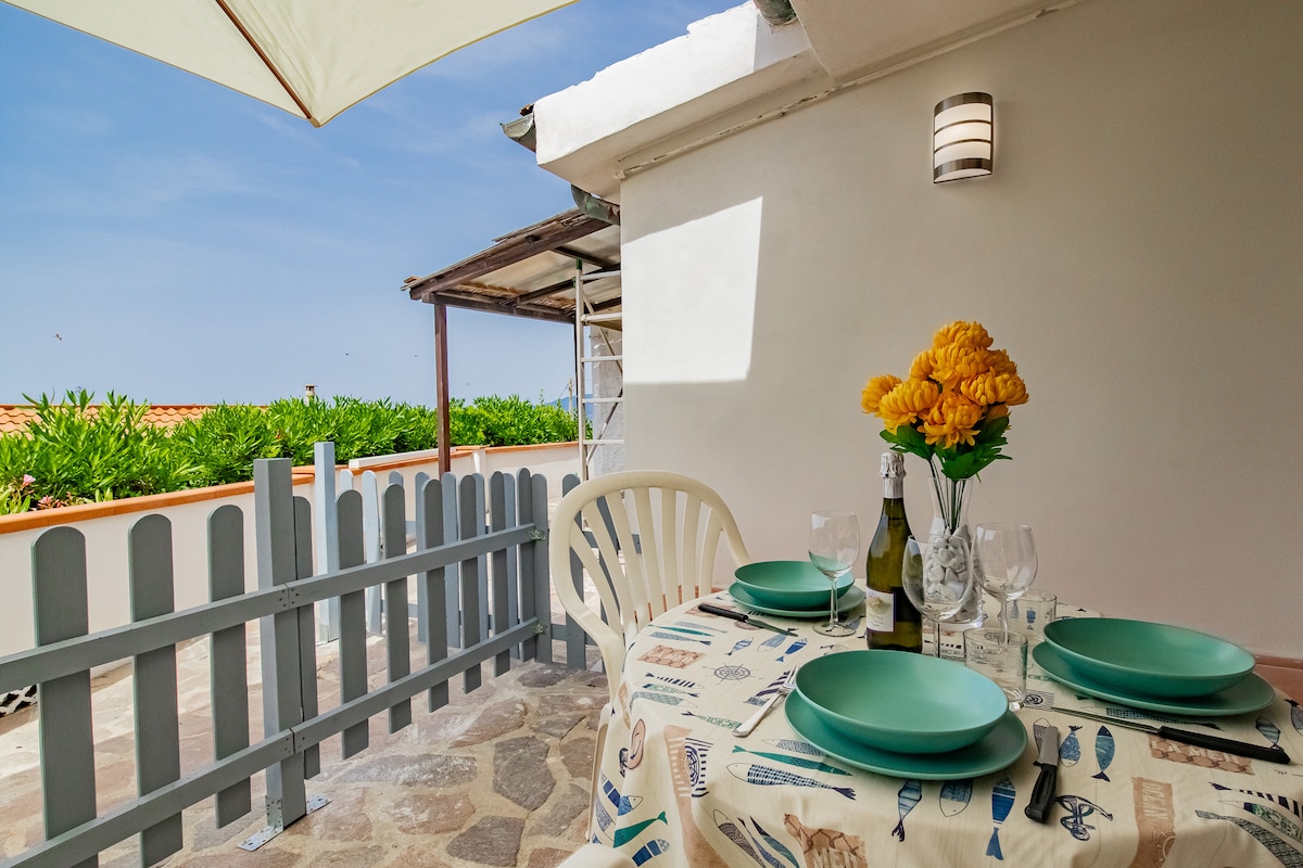 An outdoor dining area features a round table set with green plates and glassware. A vase of yellow flowers adds a pop of color. A large umbrella provides shade, and a wooden fence encloses the space, creating a private atmosphere with views of greenery.