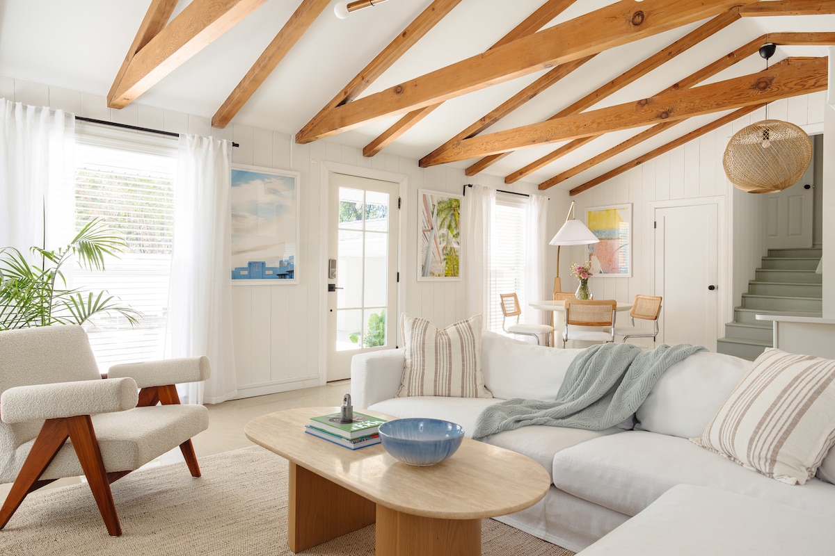 The living area features exposed wooden beams and large windows that fill the space with natural light. A comfortable sectional sofa is accented by a light throw and decorative pillows. A modern coffee table sits in the center, alongside a stylish armchair, with a dining area visible in the background.
